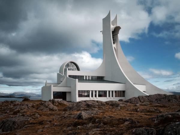 A striking, white modernist church with a tall, curved spire sits on a rocky coastal landscape under a dramatic sky.