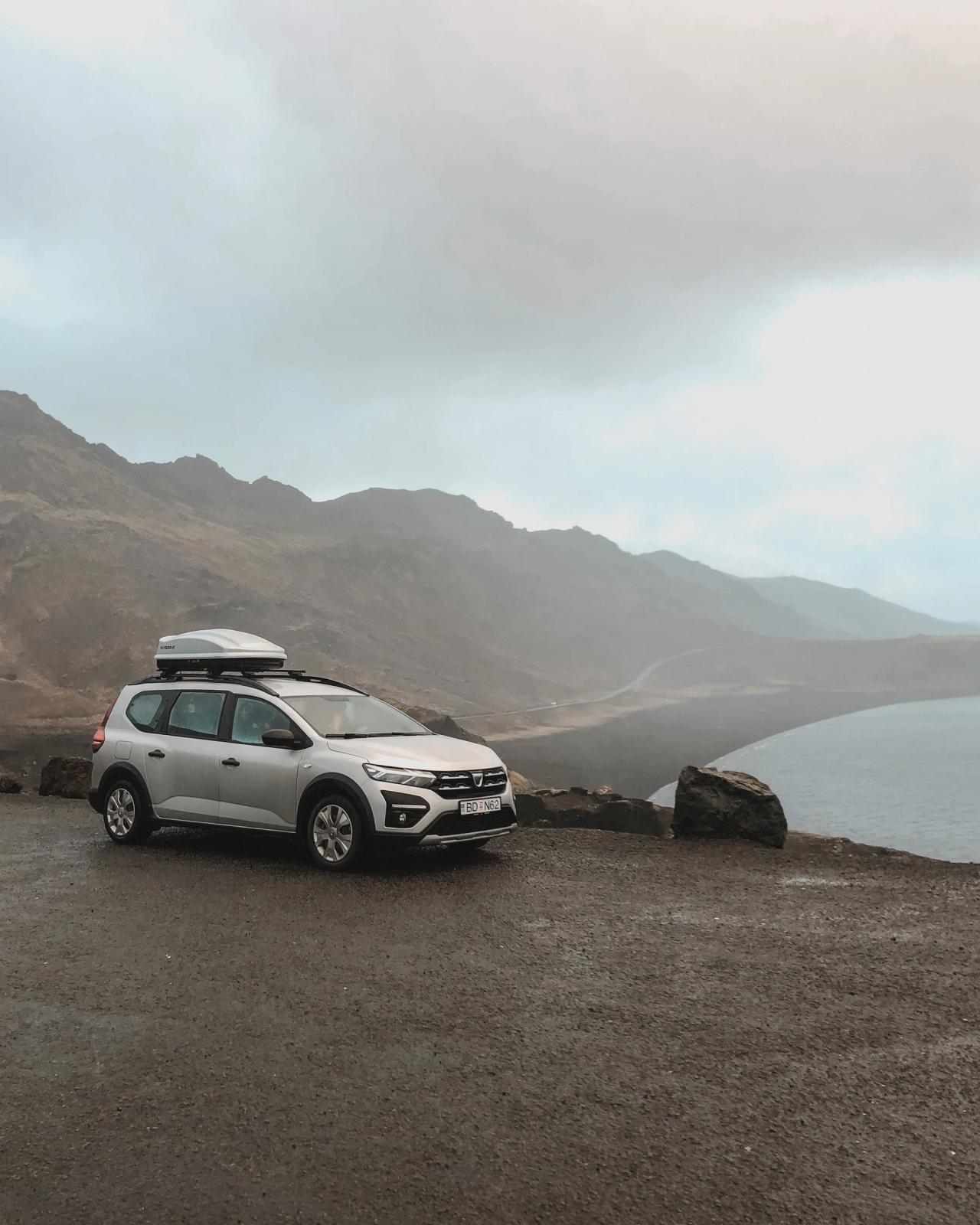 Coche plateado con cofre en el techo, aparcado en un mirador mojado sobre una bahía oscura y escarpada bajo un cielo nublado.