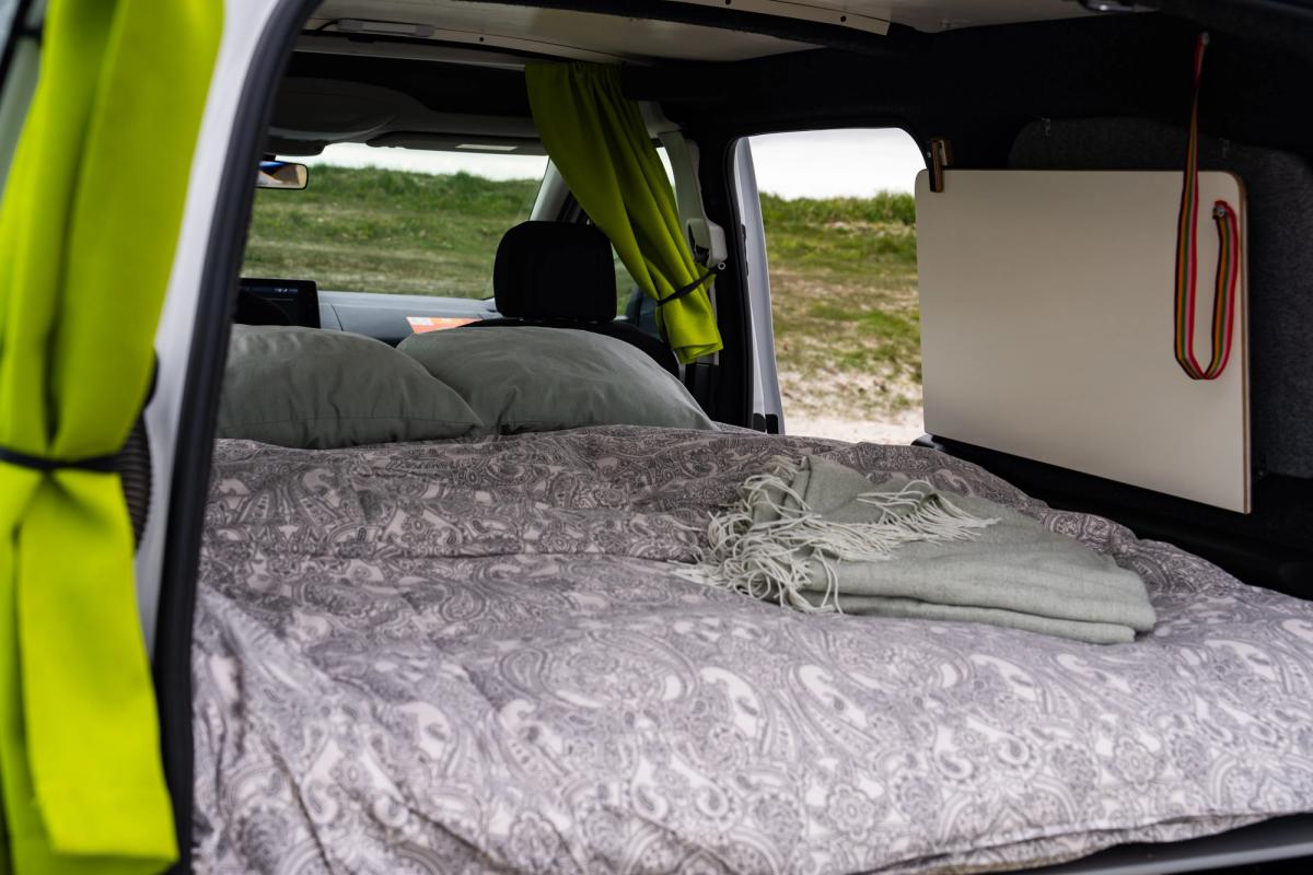 Cama hecha con edredón, almohadas y manta doblada, con un panel de madera guardado en la pared lateral