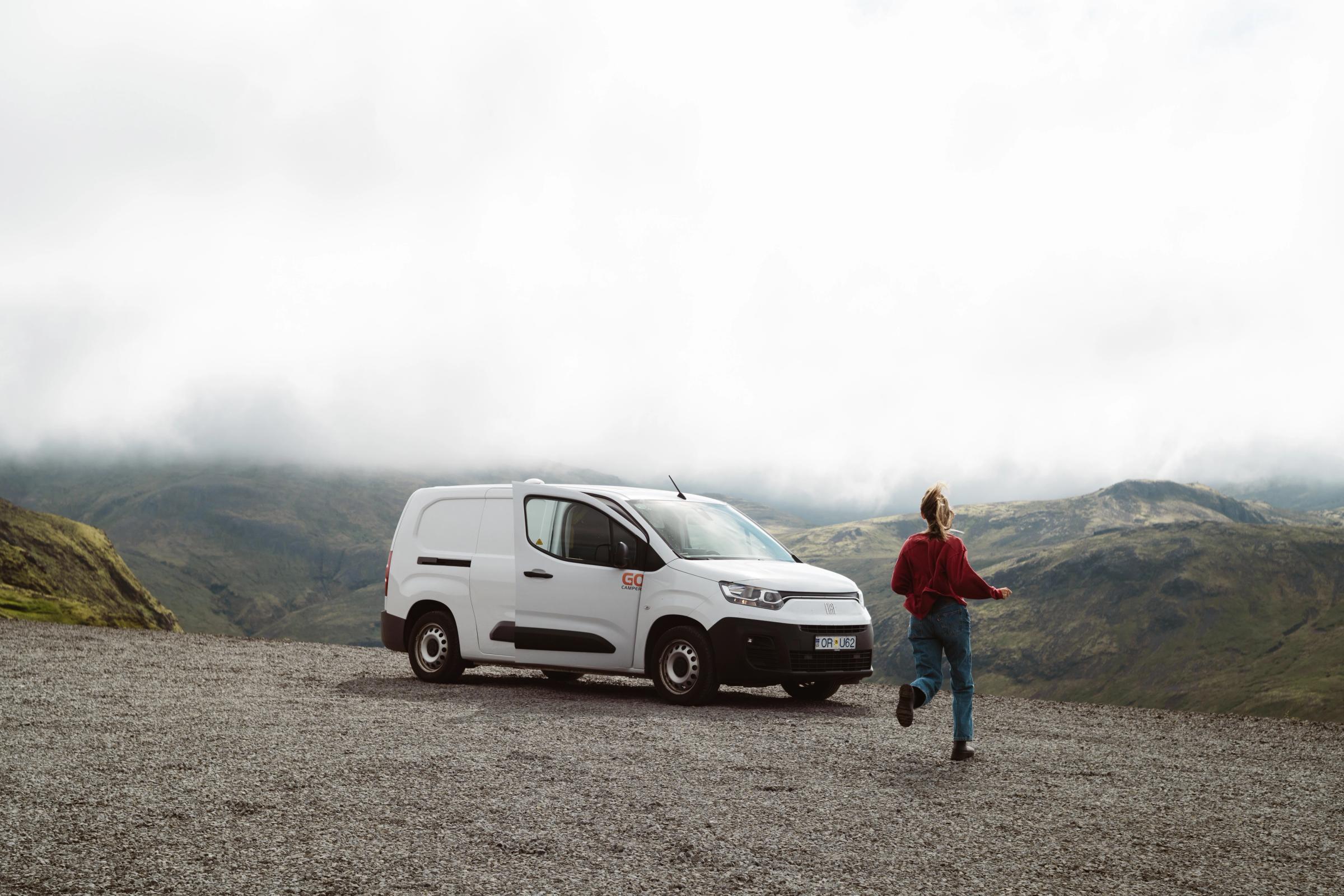 A white van parked on a gravel lot, with a person running nearby, overlooks misty green mountains.