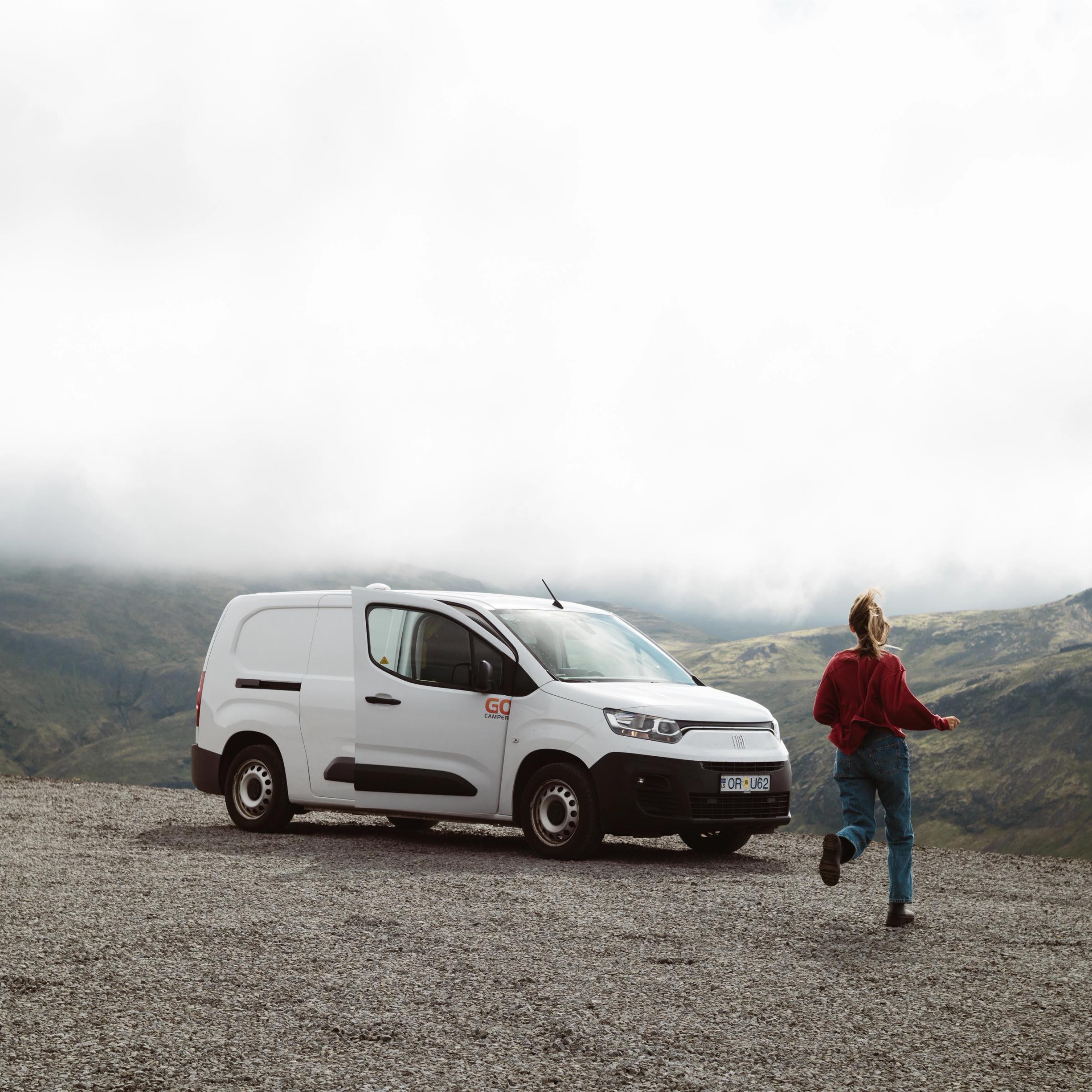 A person runs from a white commercial van on a gravel road overlooking cloudy mountains.