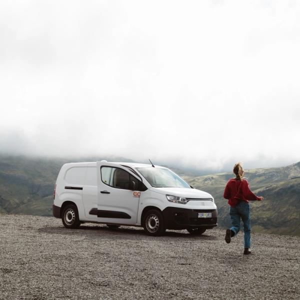 A person runs from a white commercial van on a gravel road overlooking cloudy mountains.
