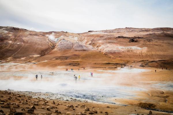 A group of people walk through a steamy, barren geothermal field with reddish-brown hills in the background.
