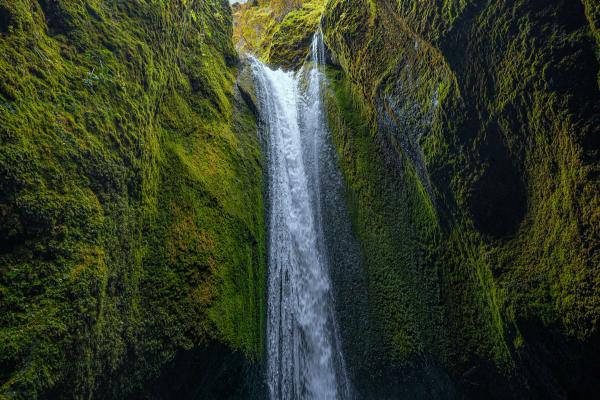 A waterfall cascades down a narrow, moss-covered canyon with a bright blue sky overhead.