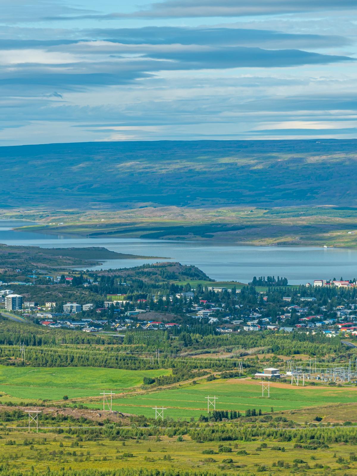 Aerial view of a town by a large lake, surrounded by green fields and distant mountains under a cloudy sky.