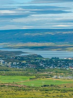 Aerial view of a town nestled between green fields and a large lake, with distant hills under a cloudy sky.