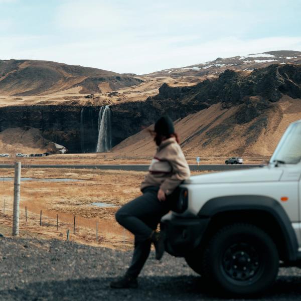 A person leans on a white SUV, looking at a waterfall in a brown mountainous landscape.