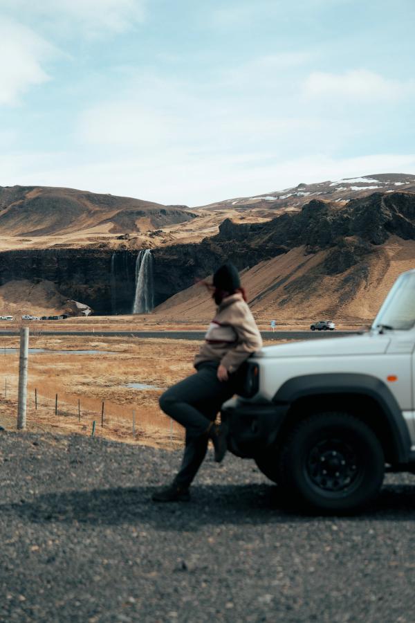 Una persona se apoya en un SUV blanco mientras contempla una cascada en un vasto paisaje montañoso de tonos marrones.