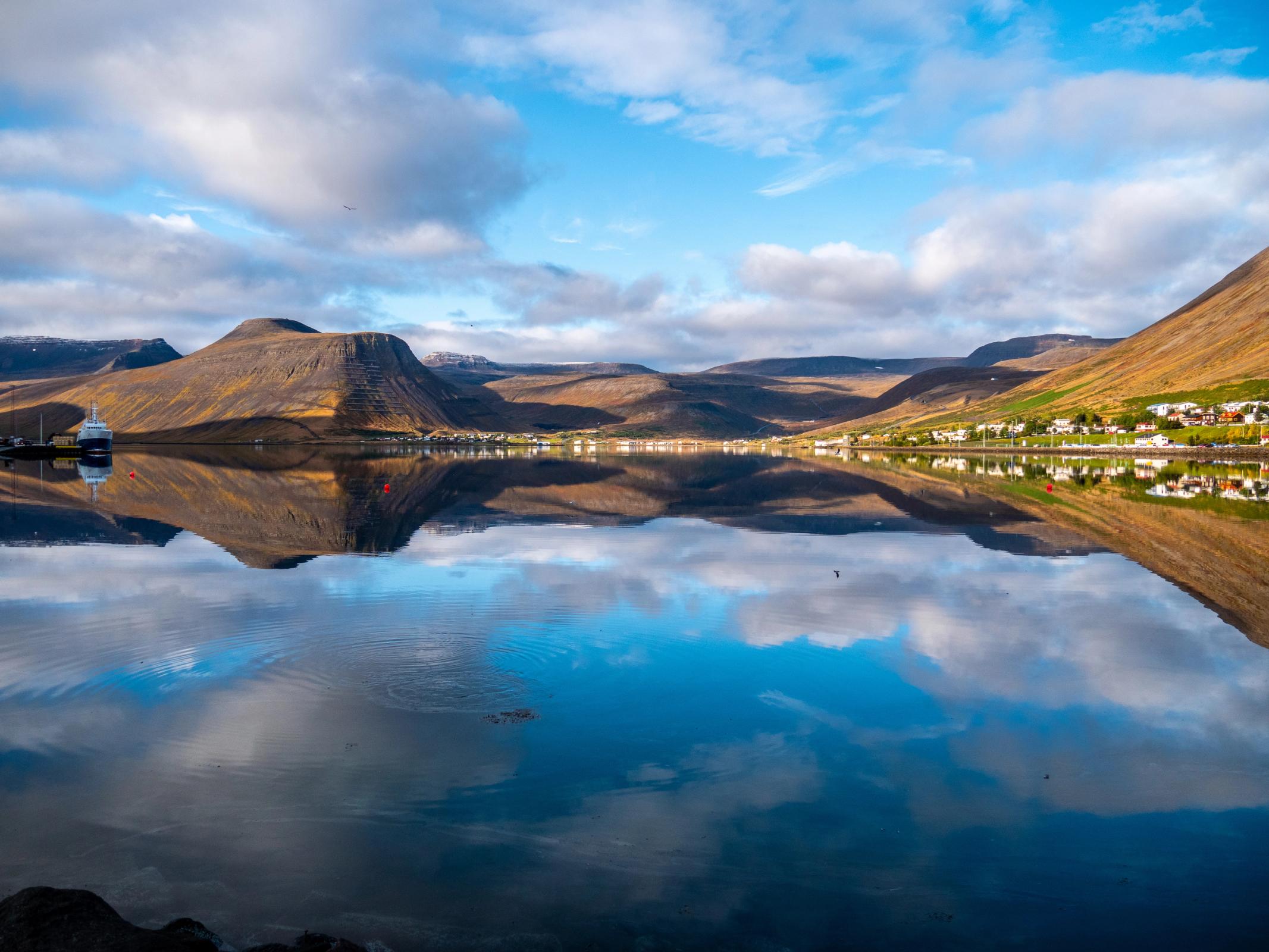 A scenic fjord landscape with mountains, a village, and a docked boat reflected in still water under a cloudy sky.