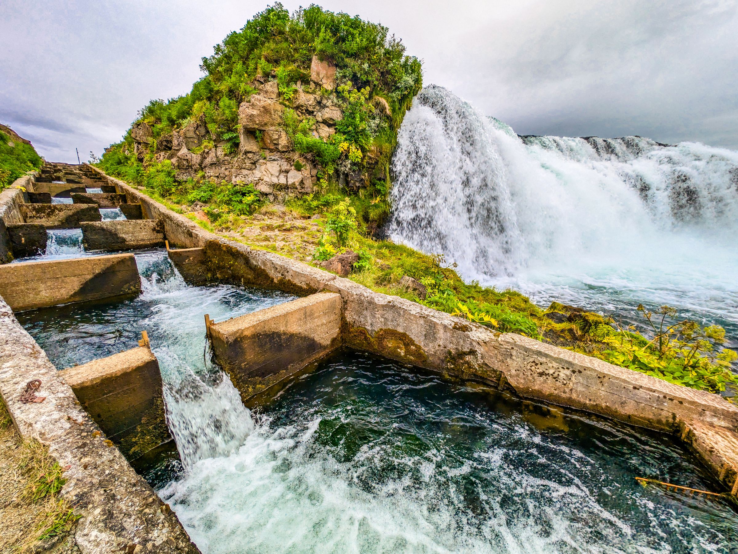 A concrete fish ladder with flowing water next to a powerful natural waterfall and green rocky cliffs.