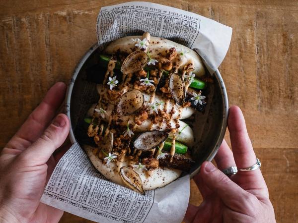 Hands hold a bowl of steamed buns topped with meat, vegetables, sauce, and small white flowers, served over newspaper on a wooden table.