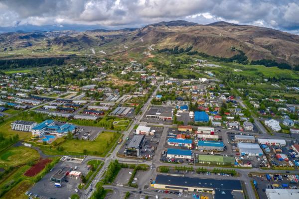 Aerial view of a town nestled in a valley with buildings, green areas, and roads, backed by mountains with visible steam.