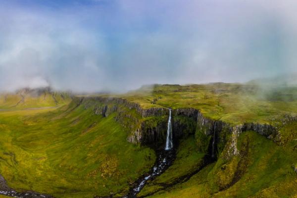 Panoramic view of a waterfall plunging from a misty, green cliff into a winding river and a bay.