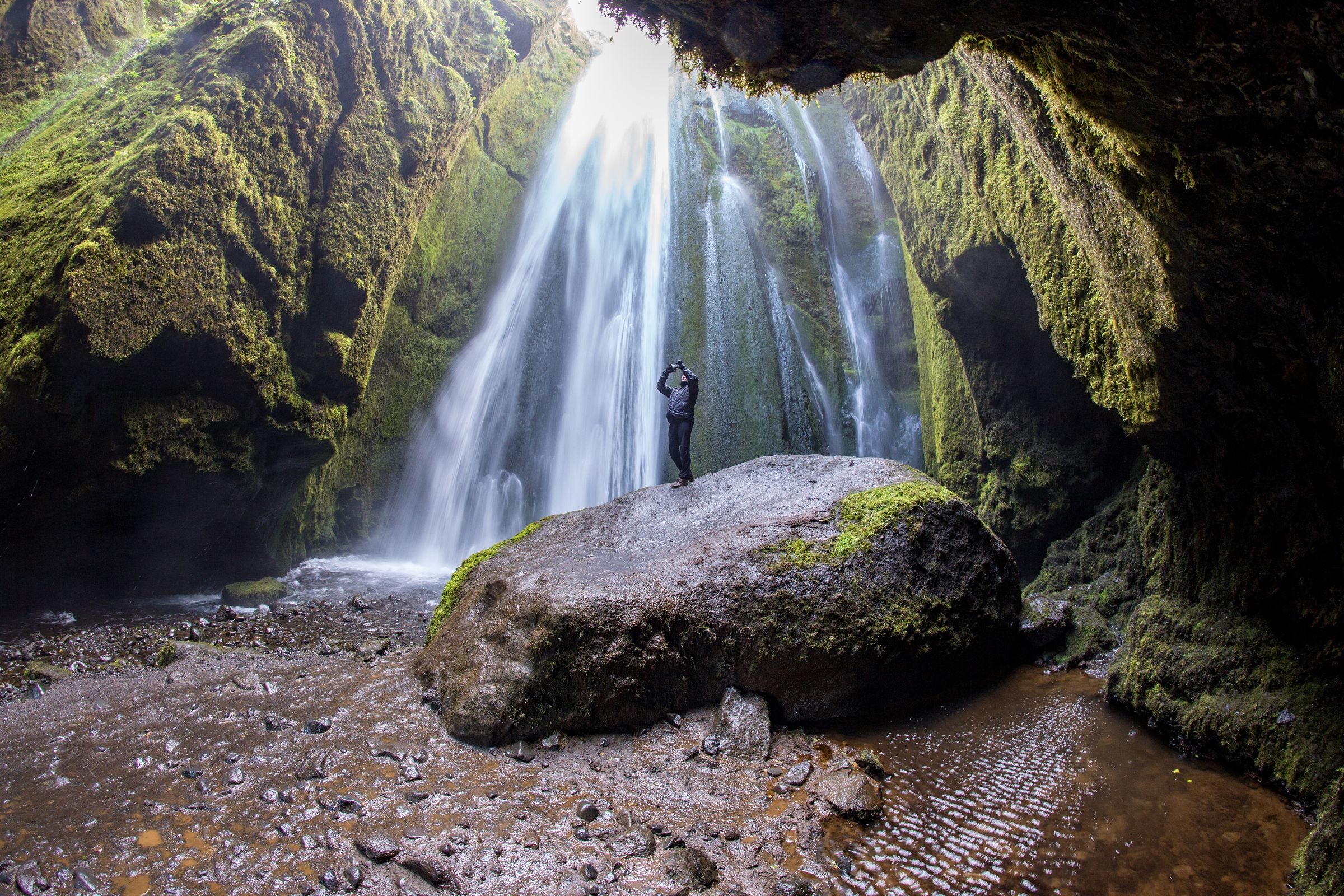 A person stands on a large rock inside a moss-covered grotto, facing a tall waterfall.