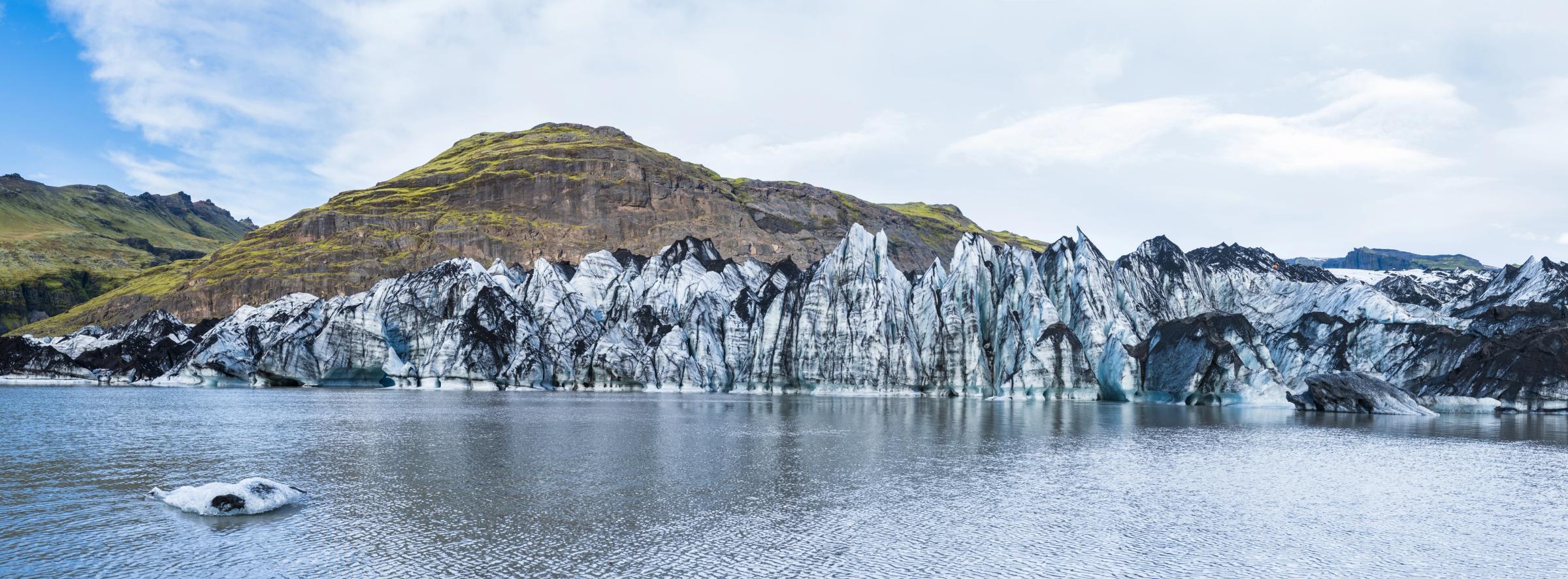 Jagged, ash-streaked glacier in a lagoon with green mountains behind it.
