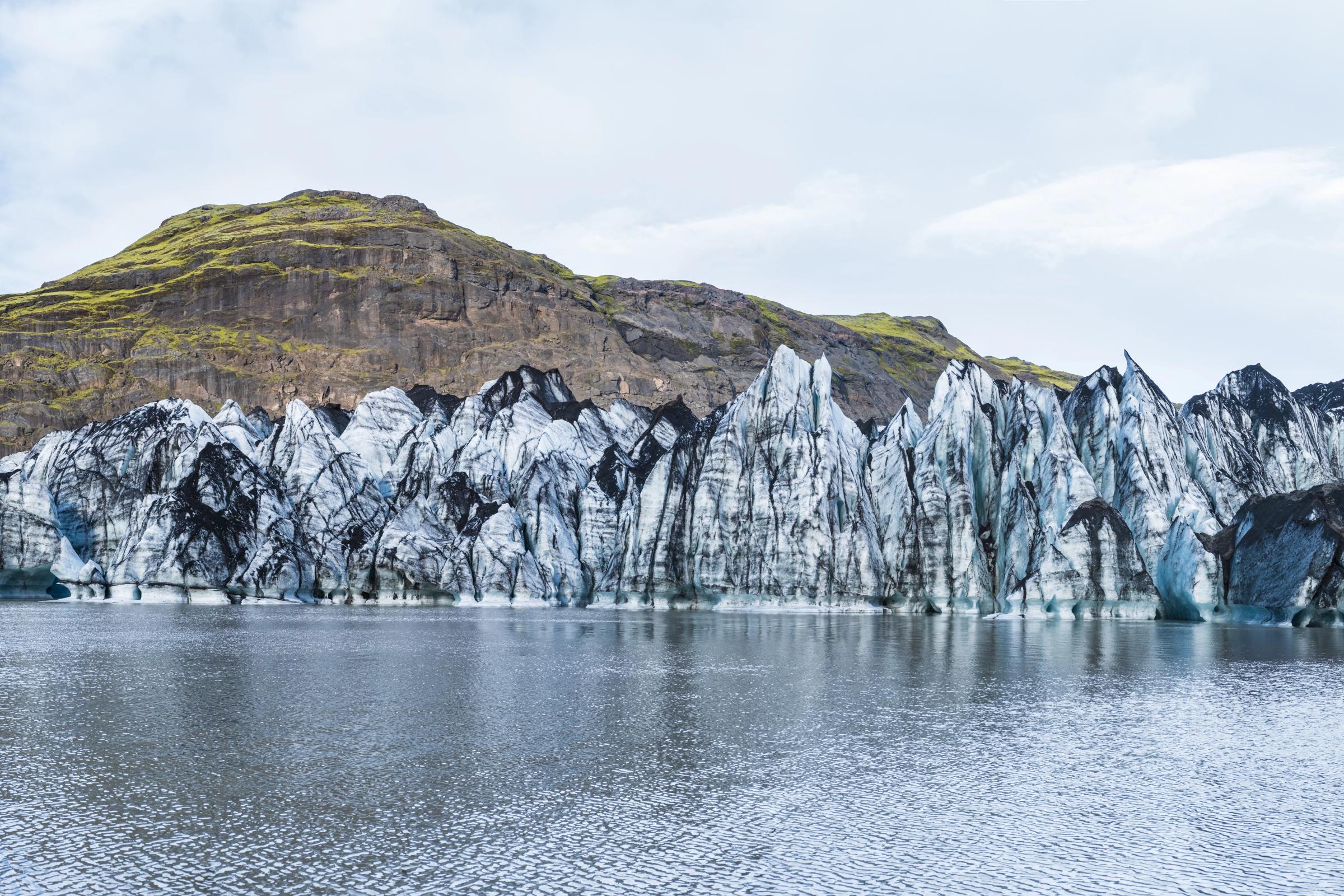 Jagged, ash-streaked glacier in a lagoon with green mountains behind it.