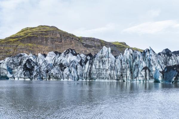 Jagged, ash-streaked glacier in a lagoon with green mountains behind it.
