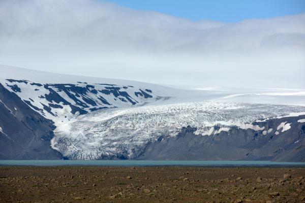 A glacier flows into a light blue lake, flanked by dark rocky mountains and a barren foreground.