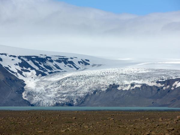 A large glacier extends into a turquoise lake, with barren rocky ground in the foreground and a cloudy sky.