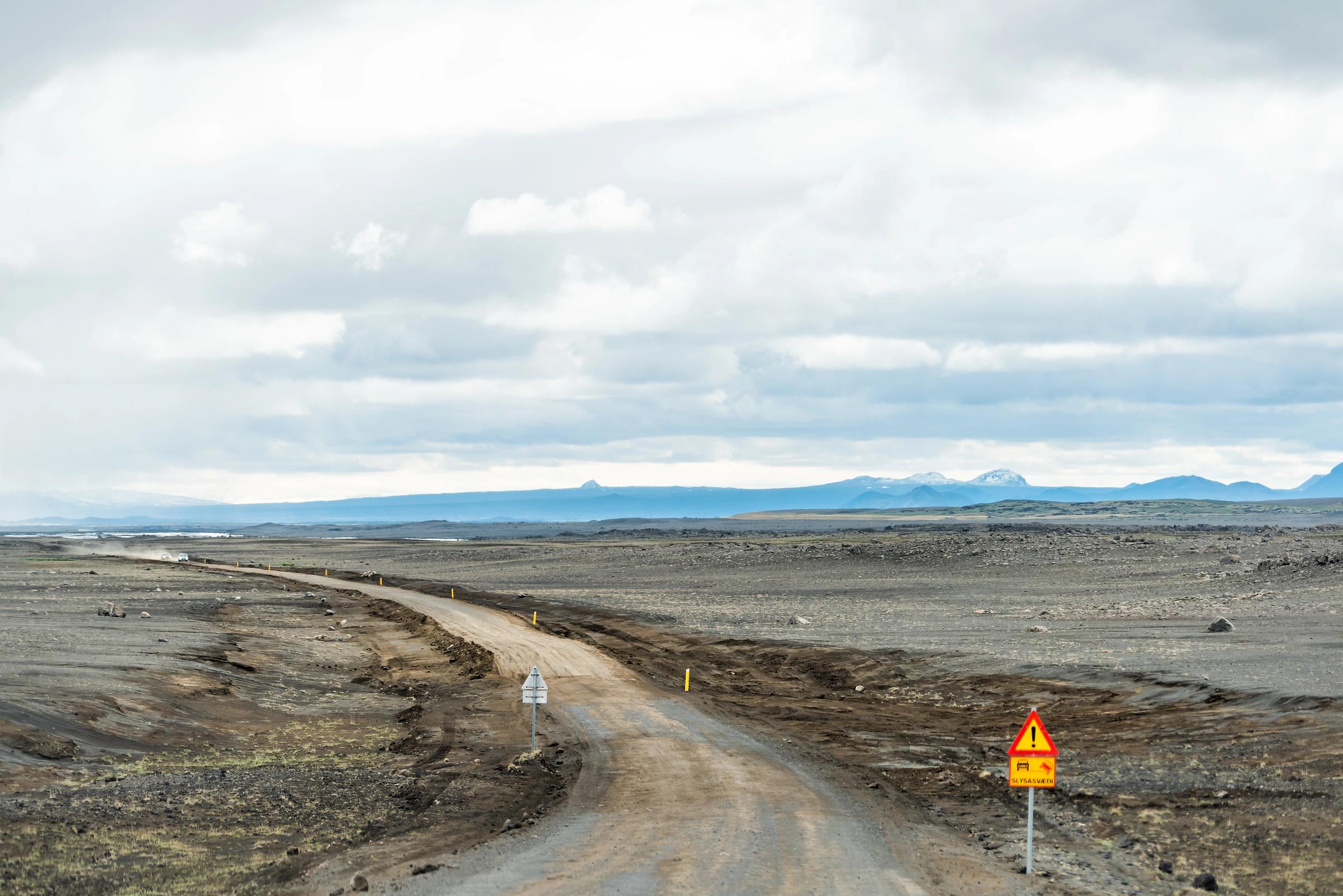 A dirt road winds through a desolate, rocky landscape under a cloudy sky, with a warning sign on the right.