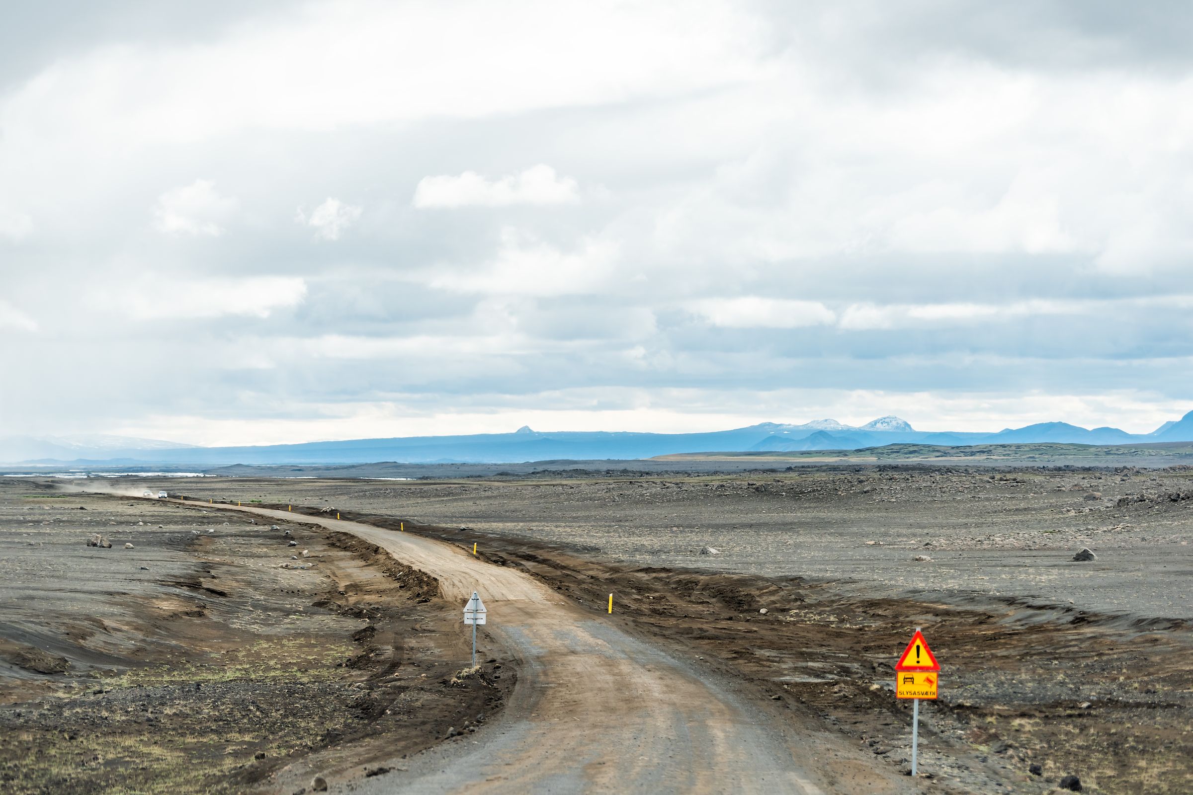 A dirt road winds through a desolate, rocky landscape under a cloudy sky, with a warning sign on the right.