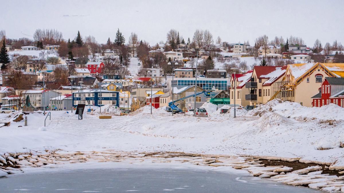 A winter scene shows a town with colorful buildings on a snow-covered hillside overlooking an icy bay.