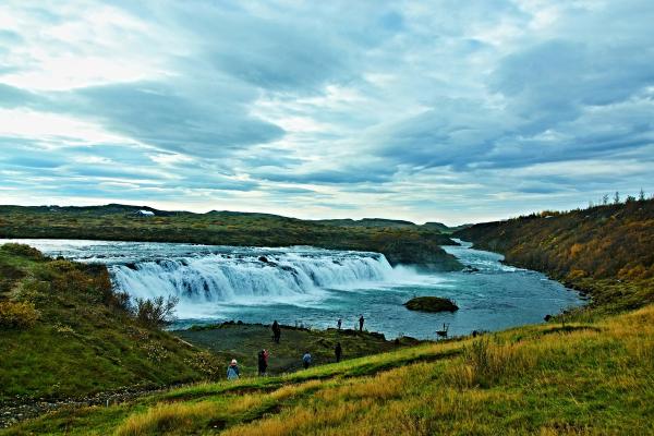 A wide waterfall in a natural landscape with green and autumn-hued hills under a cloudy sky.