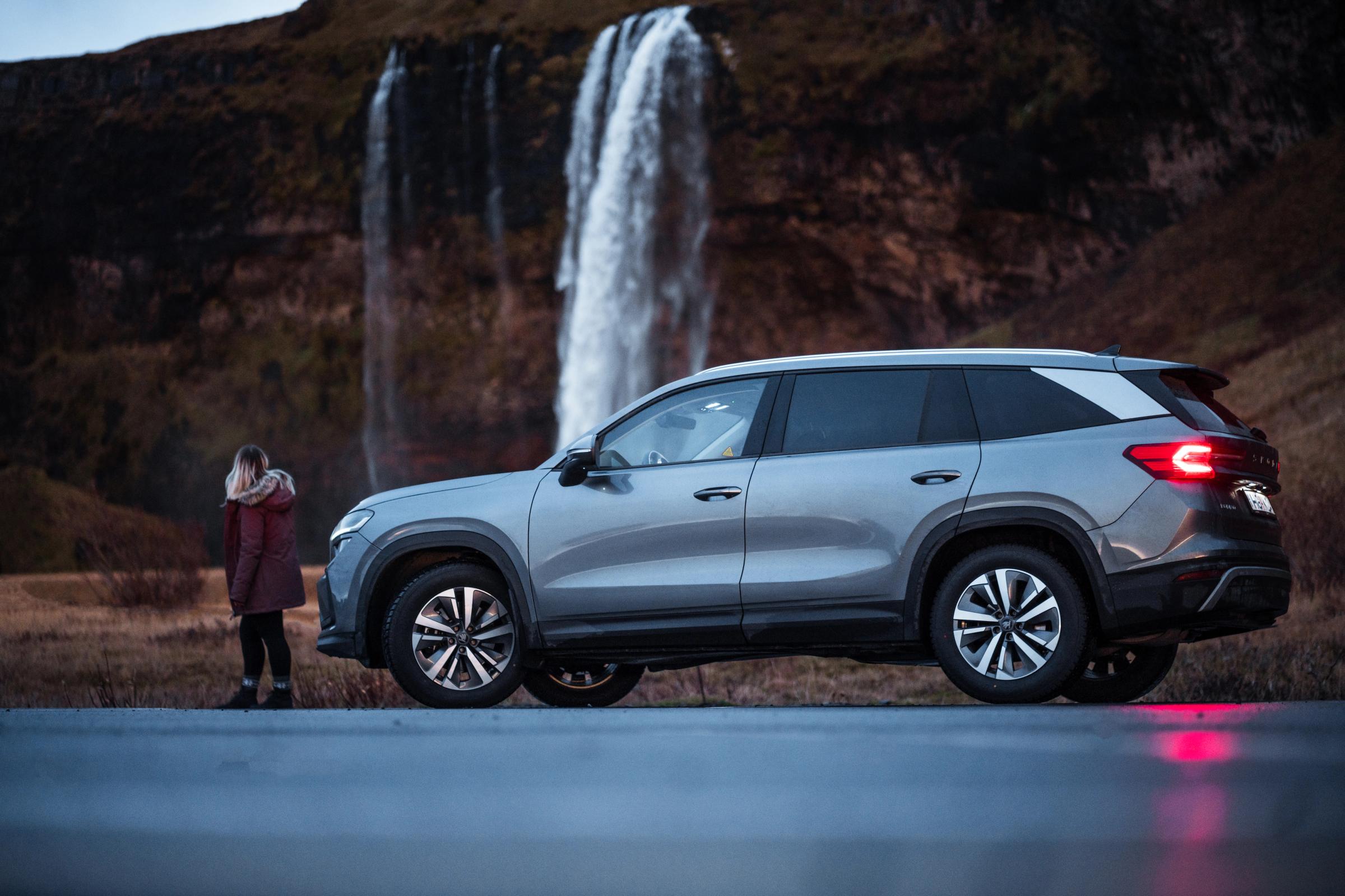 Vista lateral do Skoda Kodiaq junto a uma cascata Skoda Kodiaq visto de lado, estacionado junto a uma cascata, com uma pessoa por perto