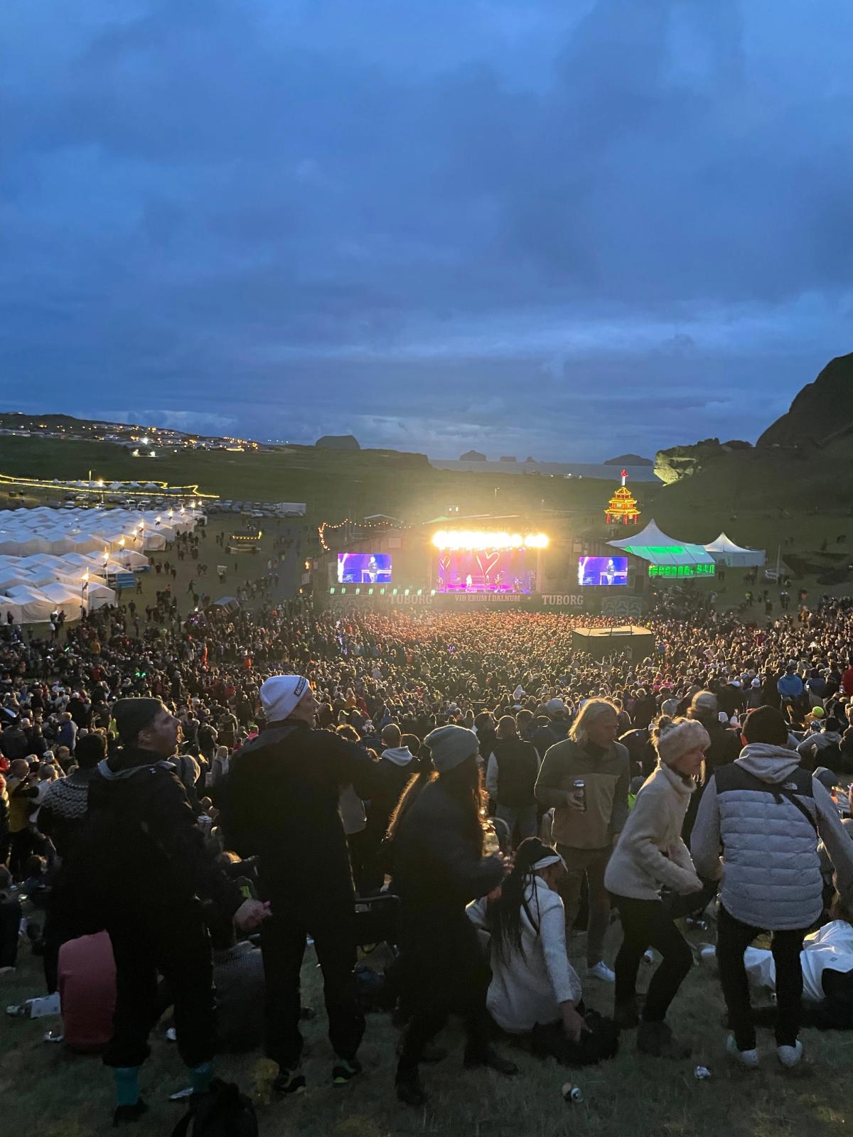 A large outdoor music festival at dusk, with a bright stage illuminating a huge crowd in a hilly landscape under a cloudy sky.