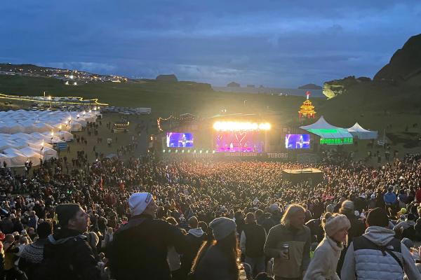 A large outdoor music festival at dusk, with a bright stage illuminating a huge crowd in a hilly landscape under a cloudy sky.