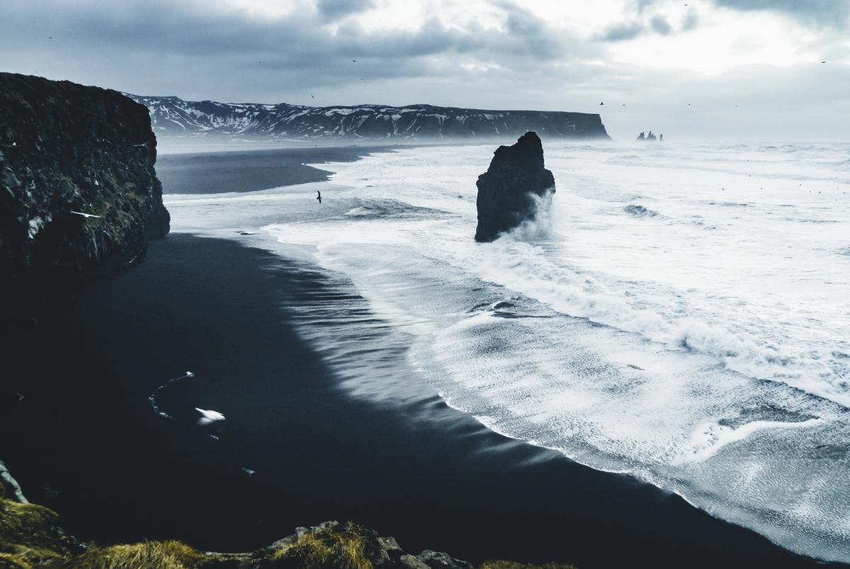 Black sand beach with white waves crashing around a prominent sea stack, cliffs, and a distant person under a cloudy sky.