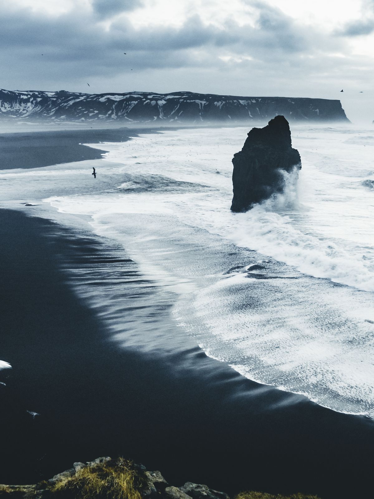 Black sand beach with white waves crashing around a prominent sea stack, cliffs, and a distant person under a cloudy sky.