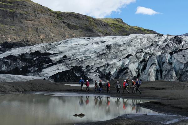 A group of hikers walks on dark ground beside a glacial pond, with a massive grey and black glacier and rocky mountains in the background.