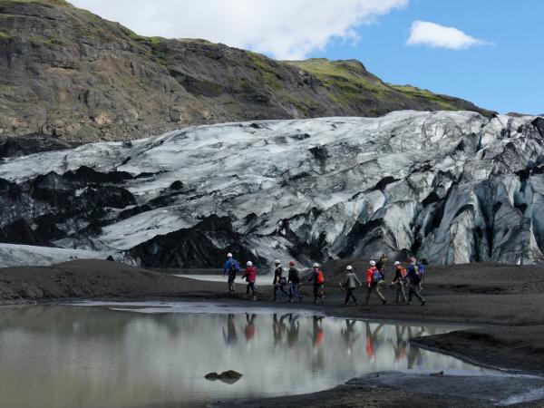 A group of hikers with helmets walk along a dark shore next to a glacial lake, with a massive, ash-covered glacier and rocky mountains beyond.