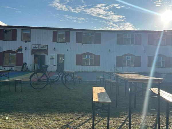 White building with red window shutters, outdoor tables and benches, a bicycle, and a rainbow flag under bright sunlight.