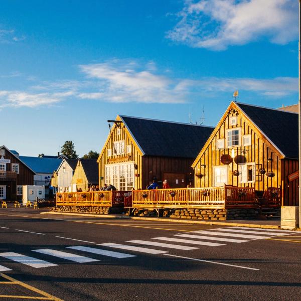 A street with a crosswalk leading to a row of rustic wooden buildings bathed in warm sunlight under a blue sky.