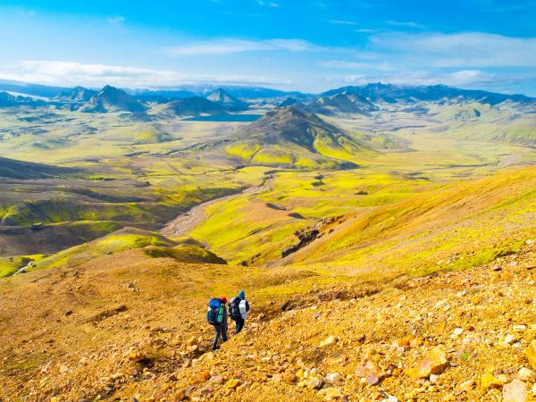 Two hikers ascend a rocky, golden slope with vibrant green and yellow valleys and distant mountains under a blue sky.
