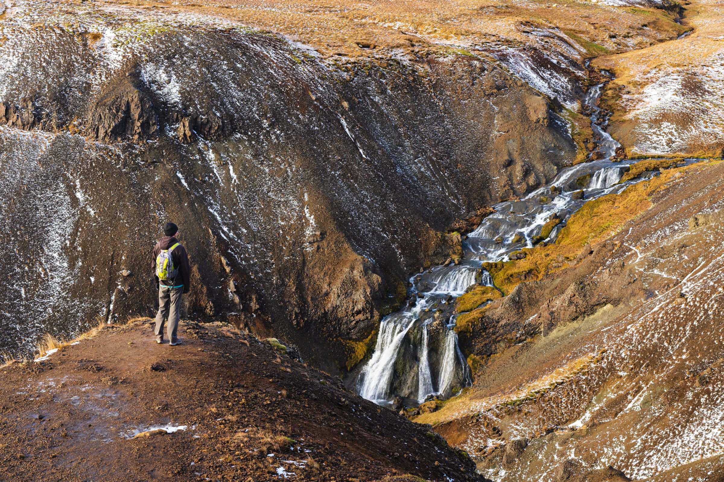 A person with a backpack stands on a rocky outcrop, overlooking a tiered waterfall cascading through a snowy, brown and yellow landscape.