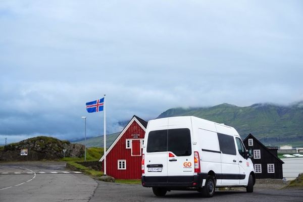 Camper blanca aparcada en frente de una casa roja con una bandera islandesa