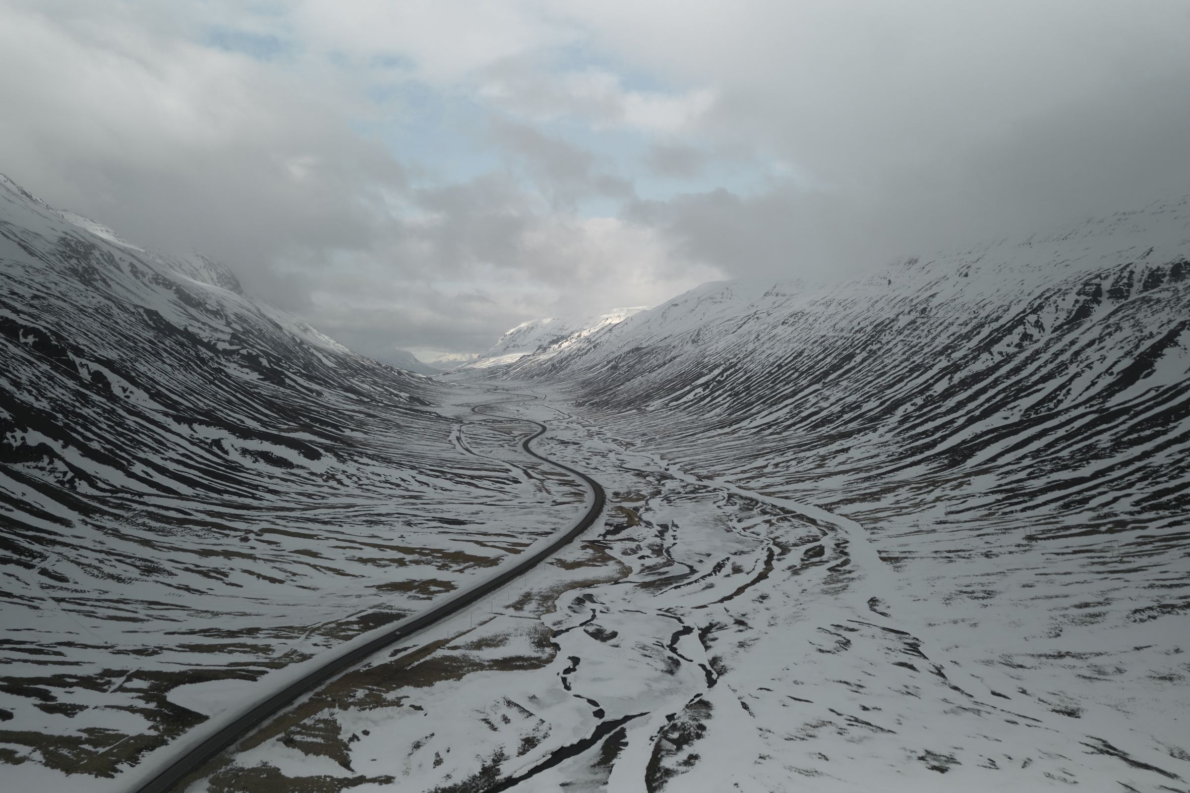 A winding road cuts through a snow-covered mountain valley under a cloudy sky.