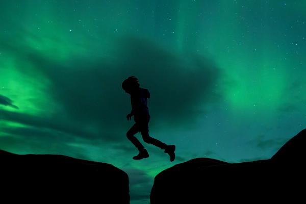 Silhouette of a person jumping a gap between dark landmasses under a green aurora sky.