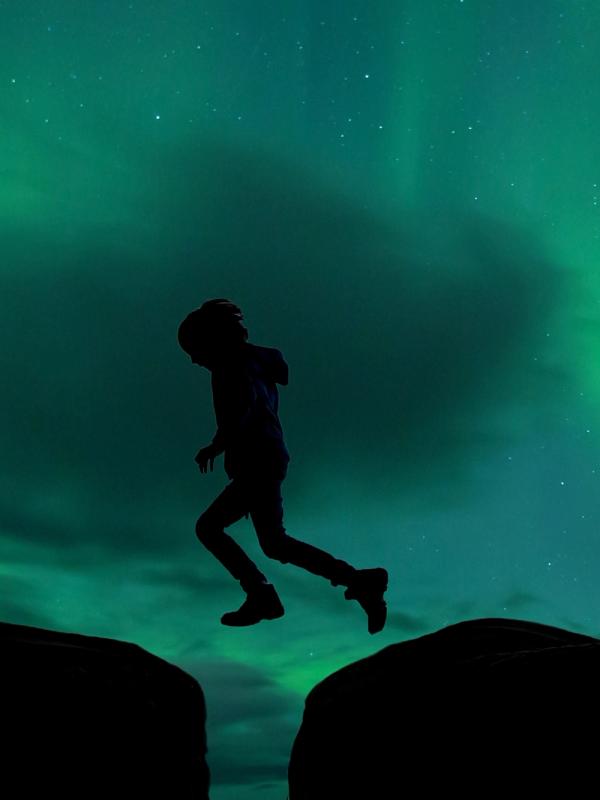 Silhouette of a person jumping a gap between dark landmasses under a green aurora sky.