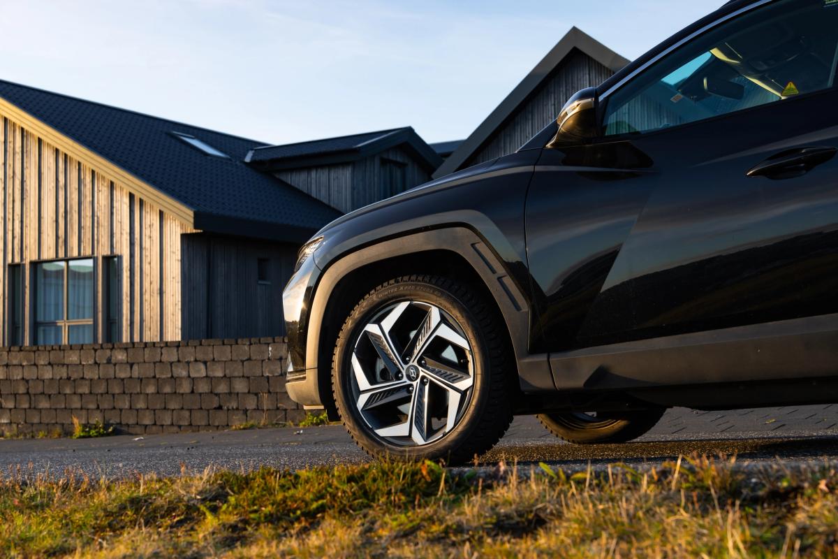 Passage de roue avant et jante alliage bicolore d’un Hyundai Tucson noir, vus en contre-plongée devant des maisons bardées de bois