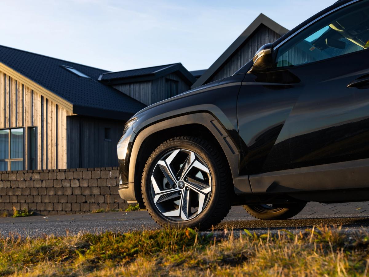 Détail de la roue avant du Tucson Passage de roue avant et jante alliage bicolore d’un Hyundai Tucson noir, vus en contre-plongée devant des maisons bardées de bois