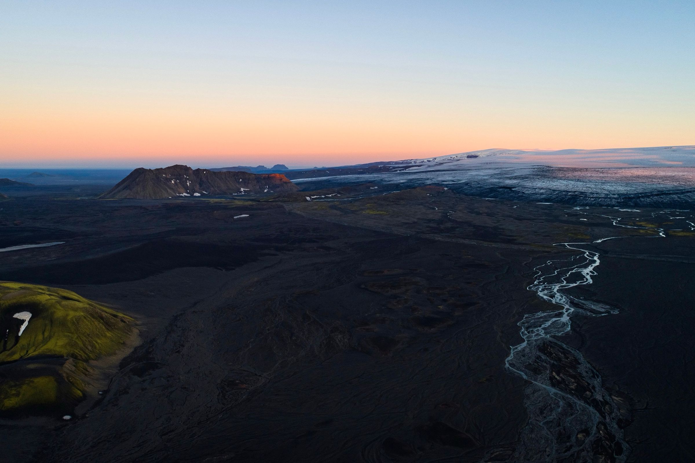 Paisaje volcánico oscuro con un río serpenteante, colinas cubiertas de musgo verde y un glaciar lejano bajo un cielo lleno de color.
