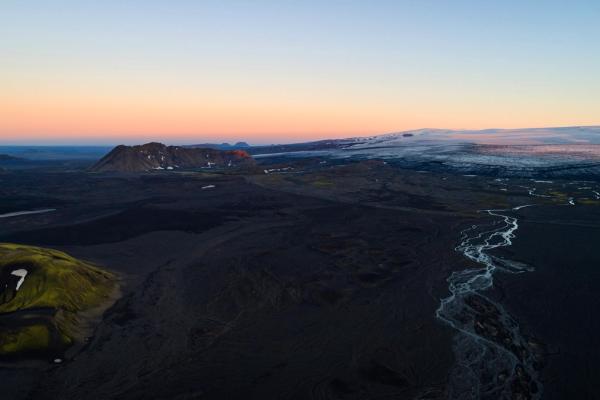 Dark volcanic landscape with a winding river, mossy green hills, and distant glacier under a colorful sky.