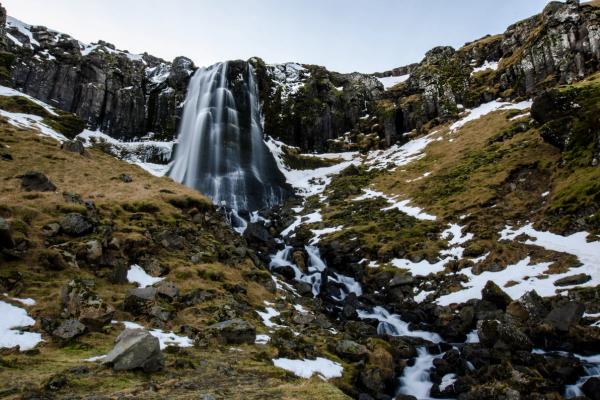 A waterfall streams down a rocky, snow-dusted slope with patches of brown and green grass.