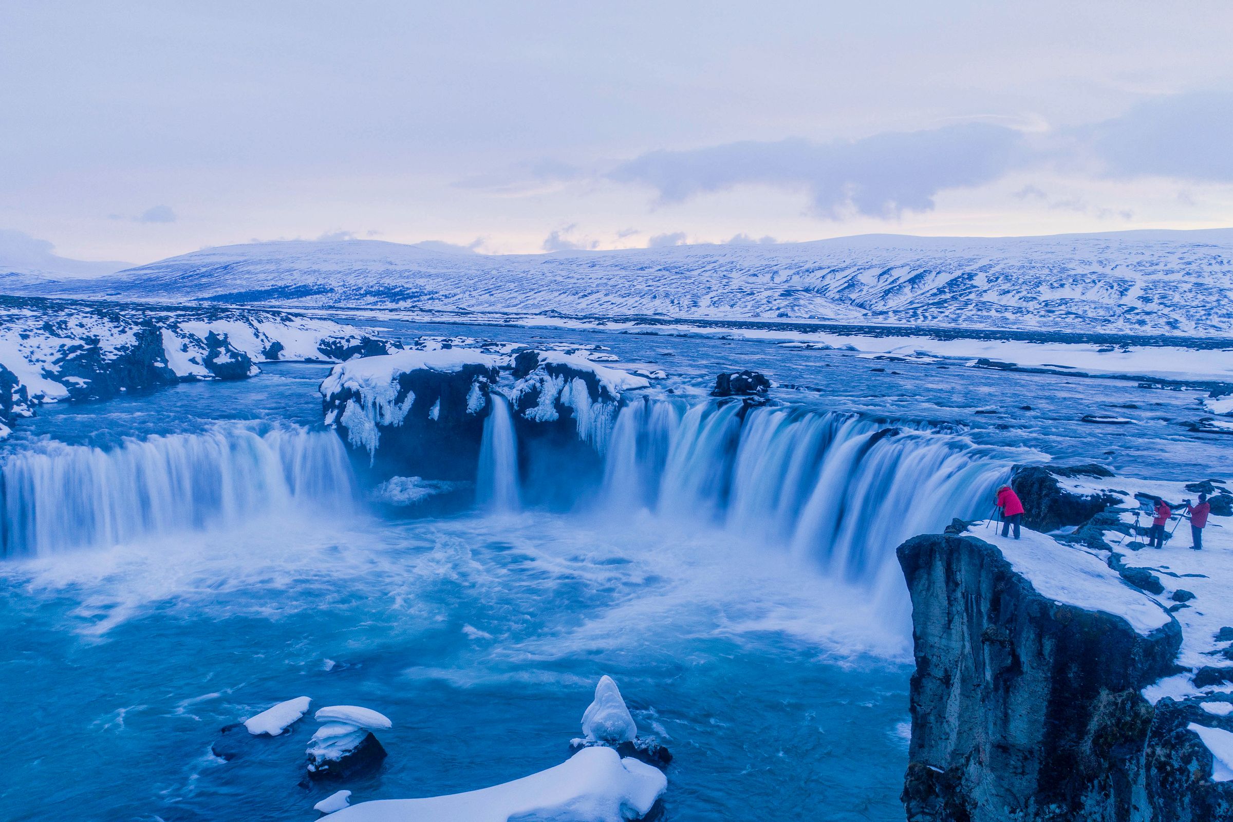 A powerful, icy blue waterfall plunges through a snowy winter landscape, with a few people on a snow-covered cliff.