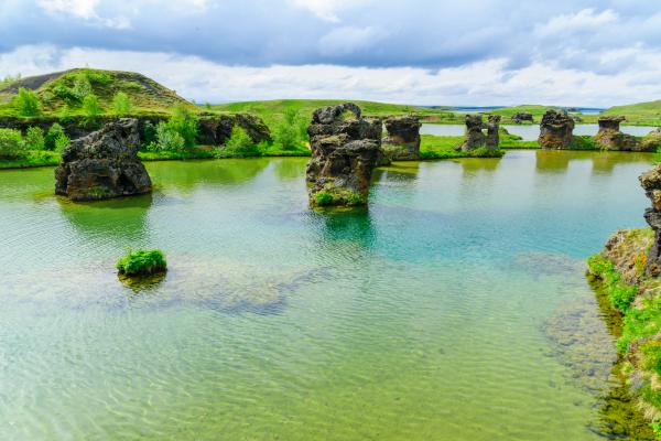 Clear green lake with dark volcanic rock pillars rising from the water, surrounded by grassy hills under a cloudy sky.