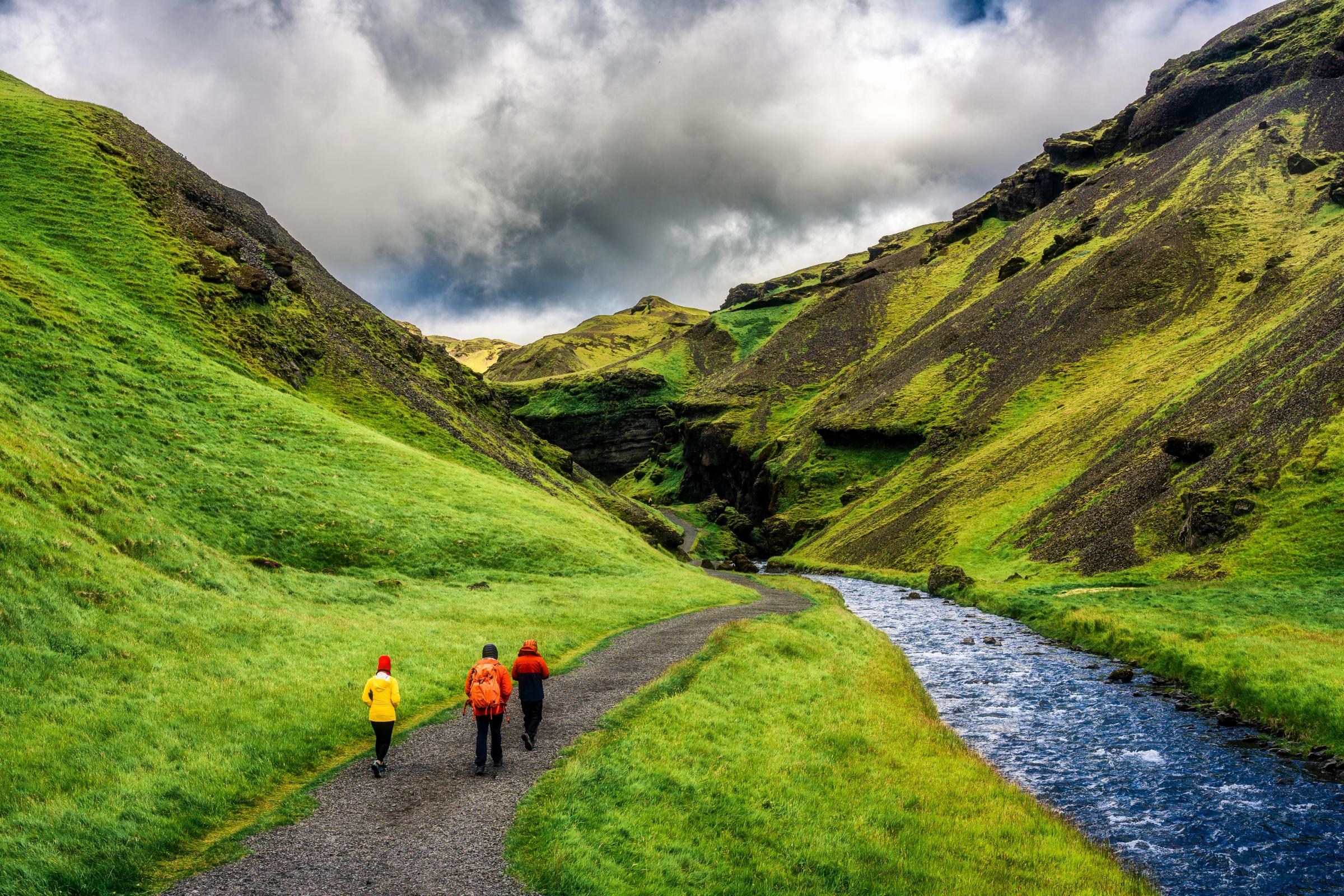 Three people hike a trail in a vibrant green valley with a river.